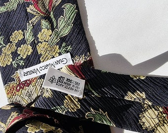 Op de afbeelding: Een zwarte zijden stropdas met een bloemenpatroon in tinten geel, rood en groen. De stropdas heeft een wit label met de tekst "Gianfranco Venturi" en een ander wit label met de tekst "100% Zijde Gemaakt in Italië Alleen chemisch reinigen".