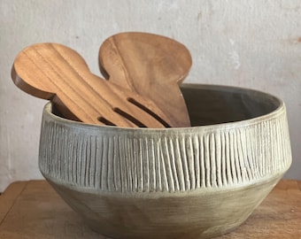 May include: A ceramic salad bowl with a textured, vertical-line design on the upper portion. The bowl is two-toned, with a lighter color on the textured top and a darker, earthy tone on the bottom. Wooden salad servers rest inside.