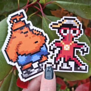 May include: Two pixel art stickers. One is an orange character with a blue shirt. The other is a red character wearing a hat and gold chain. Both stickers have a white border and are on a background of green and red leaves.