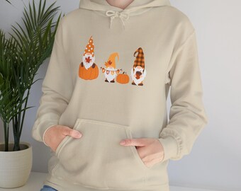 May include: Beige hoodie featuring a front graphic of three fall gnomes with pumpkins. The gnomes wear orange and brown hats and hold fall leaves. The hoodie has a kangaroo pocket and a drawstring hood.