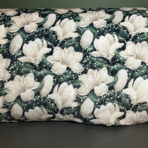 May include: A decorative pillow with a repeating pattern of large, white magnolia flowers against a dark green background. The flowers have detailed petals and are surrounded by smaller white dots and green leaves.