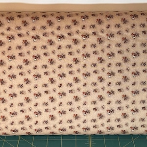 May include: A bolt of fabric featuring a light beige background with a repeating pattern of small, dark brown and reddish-brown floral clusters. The textile has a visible woven texture and is displayed horizontally across the frame.