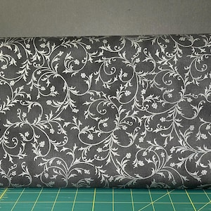May include: A roll of fabric with a dark gray background and a repeating white floral pattern. The design features swirling vines and small flower clusters. The fabric is rolled up, showing the pattern in detail.