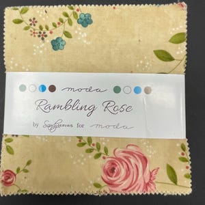 May include: A stack of fabric squares with a floral pattern, featuring pink roses, green leaves, and small blue flowers on a beige background. The label reads "Rambling Rose" by Sandy Gervais for Moda.