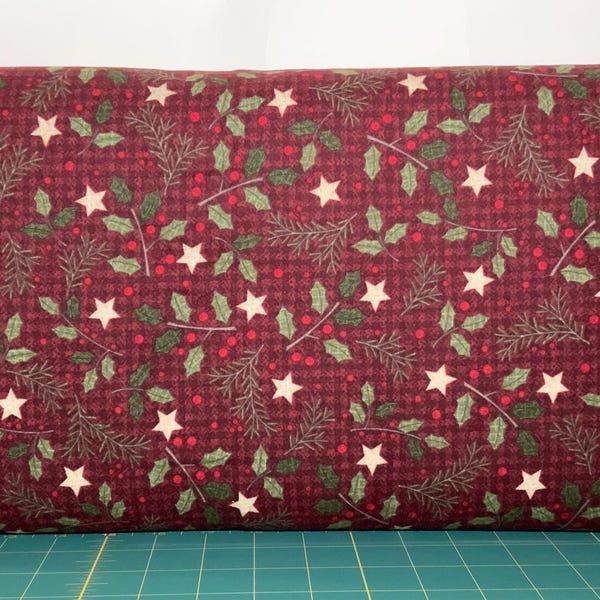 May include: A bolt of fabric featuring a dark burgundy background with a subtle black plaid pattern. The print displays a repeating holiday motif of green holly leaves, small red berries, thin pine branches, and cream-colored five-pointed stars.