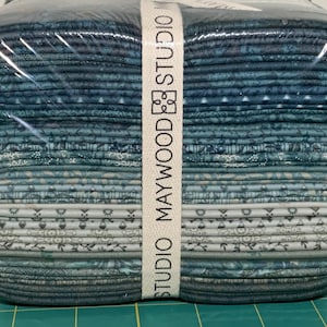 May include: A stack of folded fabric squares wrapped in clear plastic with a white paper band labeled MAYWOOD STUDIO. The fabric features a gradient of blue and gray tones, ranging from dark navy to light slate, with various small floral and geometric patterns.