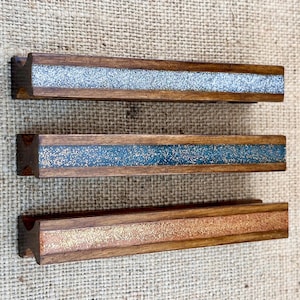 Puede incluir: Tres tiradores de cajón de madera con una incrustación decorativa de purpurina. Los tiradores son en tonos plata, azul verdoso y dorado.