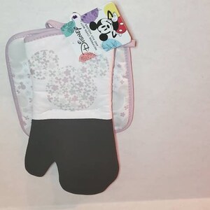 May include: A white oven mitt with a black heat-resistant lining and a Mickey Mouse silhouette design. The mitt is paired with a matching white pot holder with a pink border. The packaging includes a tag with the Disney logo and Mickey Mouse.