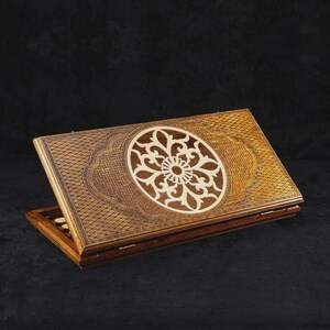May include: A wooden backgammon set with intricate carvings. The top of the game board features a central floral design in a lighter wood, surrounded by detailed patterns. The set is open, revealing the game pieces.