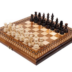 May include: A wooden chess set with a decorative inlaid border. The chessboard features light and dark squares, with black and white chess pieces arranged for play. The set is displayed on a white background.