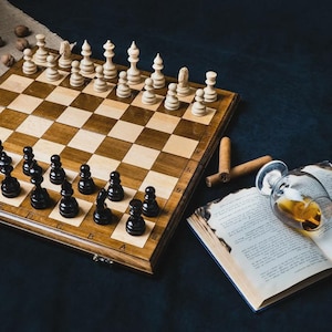 Op de afbeelding: Een houten schaakspel met zwarte en witte stukken op een dambord. Een glas amberkleurige vloeistof, sigaren en een open boek liggen naast het bord. De scène speelt zich af op een donkere ondergrond.