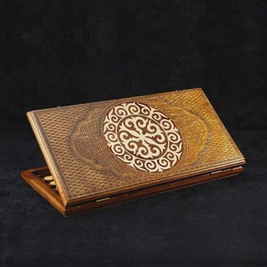 May include: A wooden backgammon set with detailed carvings. The set has a light brown exterior with a central, circular design in a lighter wood tone. The board is open, showing the playing surface and game pieces.