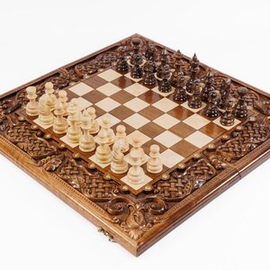 May include: A wooden chess set with intricately carved detailing around the board's perimeter. The board features a light and dark checkered pattern, with chess pieces in light and dark brown. The set is displayed on a white background.