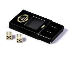 May include: Two ivory dice with black dots next to a black box with a sliding drawer. The box has a gold emblem and the word "Dice" printed on it. The dice are for a game.