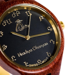 Peut inclure: Gros plan d'une montre-bracelet en bois avec un cadran sombre et des accents dorés. Le cadran de la montre comporte des chiffres et des aiguilles dorés, avec le nom "Hrachya Ohanyan" et "SWISS MOVT" imprimés. Le bracelet est en bois brun.