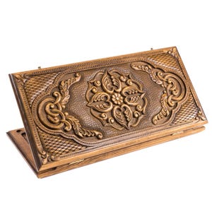 May include: A wooden backgammon board with intricate carved floral and scroll designs. The board is open, revealing the playing surface and detailed carvings. The wood has a warm, natural tone, and the craftsmanship is evident in the fine details.