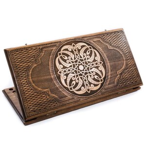 May include: A wooden backgammon board with a detailed, carved design. The board is a warm brown color with a lighter, intricate circular pattern in the center. The edges feature a diamond-patterned border.