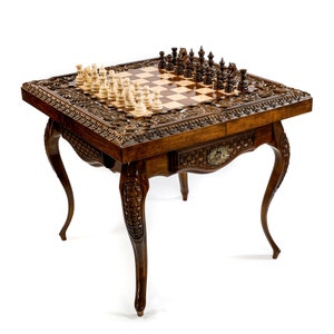 May include: A detailed wooden chess table with carved floral designs and curved legs. The chessboard is inlaid with light and dark squares, and chess pieces are arranged on the board. The table has a small drawer with a decorative emblem.