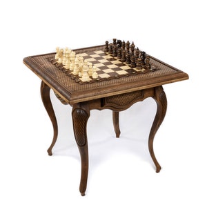 May include: A wooden chess table with a checkered playing surface and intricately carved details. The table features a drawer and curved legs. Chess pieces are arranged on the board, with light and dark pieces ready to play.