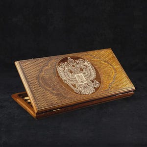 May include: A wooden backgammon set with a detailed carved design. The set features a central emblem of a double-headed eagle, with intricate patterns and a warm, natural wood finish. The board is open, revealing the playing surface and game pieces.