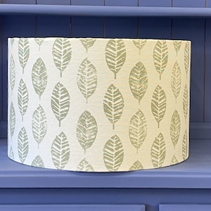 May include: A drum lampshade featuring a cream-coloured fabric with a repeating pattern of green leaf motifs. The lampshade is displayed on a blue surface.