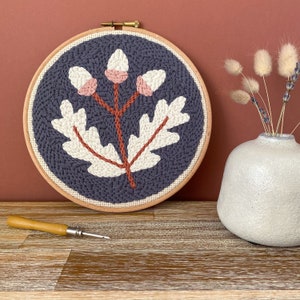 May include: A grey embroidery hoop with a white and brown acorn design. The design features three acorns with a stem and leaves. The hoop is on a wooden surface.
