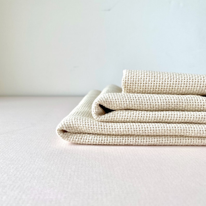 May include: A stack of three beige Aida cloth squares, folded neatly. The fabric is woven with a grid pattern, ideal for cross-stitch or needlepoint projects.