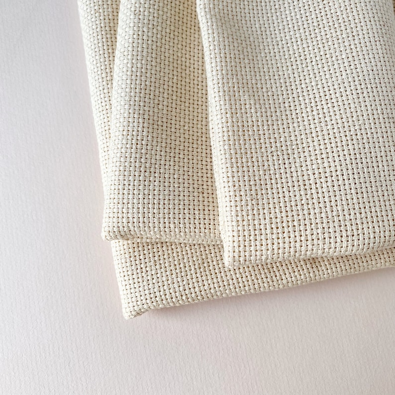 May include: Two pieces of beige Aida cloth folded together. The cloth is a woven fabric with a grid pattern, commonly used for cross-stitch embroidery.