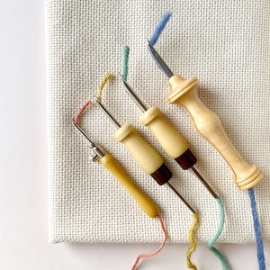 May include: Four embroidery needles with wooden handles and different colored threads. The needles are on a white canvas background.