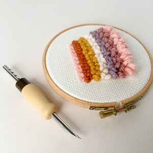 May include: A close-up of a wooden embroidery hoop with a colourful yarn rainbow design. A wooden needle tool is also pictured.