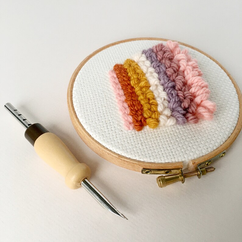 May include: A close-up of a wooden embroidery hoop with a colourful yarn rainbow design. A wooden needle tool is also pictured.