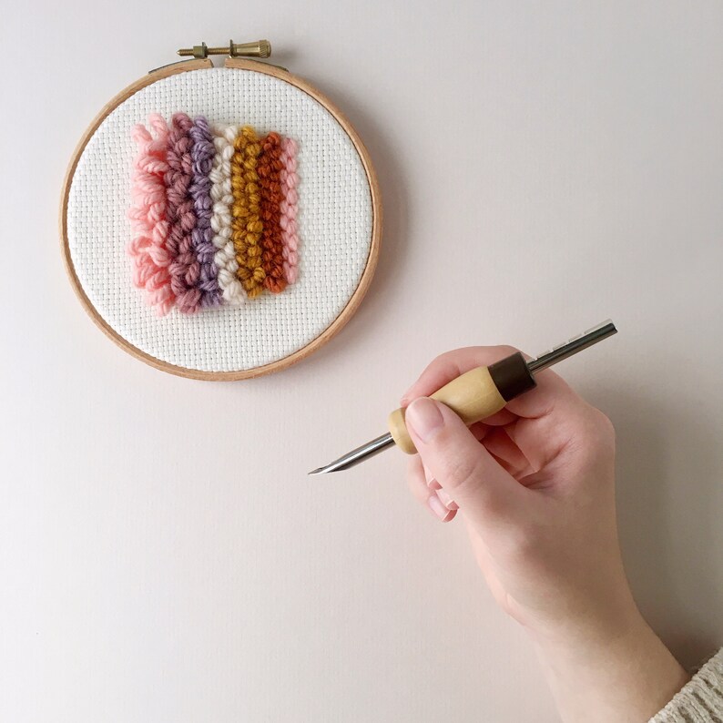 May include: A close-up of a wooden embroidery hoop with a colourful yarn tapestry inside. A hand holds a wooden needle tool for punch needle embroidery.