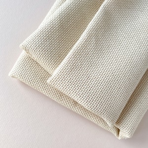 May include: Two pieces of white Aida cloth folded together. The cloth is a woven fabric with a grid pattern, commonly used for cross-stitch projects.