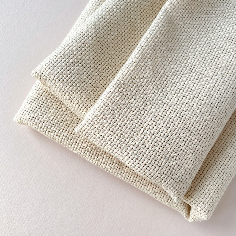 May include: Two pieces of white Aida cloth folded together. The cloth is a woven fabric with a grid pattern, commonly used for cross-stitch projects.