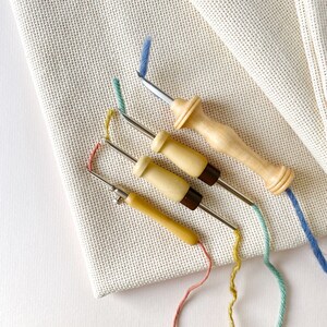 May include: Four wooden embroidery needles with metal tips and colorful threads. The needles are arranged on a piece of white Aida cloth.
