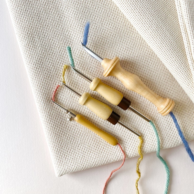 May include: Four wooden embroidery needles with metal tips and colorful threads. The needles are arranged on a piece of white Aida cloth.