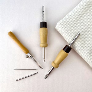 May include: Three wooden embroidery tools with metal needles. Two of the tools have a black handle and a metal needle with a small loop at the end. The third tool has a wooden handle with a metal needle and a small loop at the end. The tools are on a white surface.