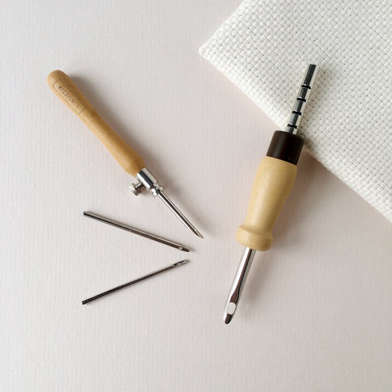 May include: Two wooden embroidery tools with metal needles. The tool on the left has a wooden handle and a metal needle with a small knob. The tool on the right has a wooden handle with a brown cap and a metal needle with a series of holes along the shaft. Three additional metal needles are lying on the table.