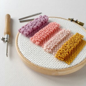 May include: A close-up of a wooden embroidery hoop with colourful yarn woven into a striped pattern. The yarn is in shades of pink, peach, and yellow. The hoop is on a white surface.