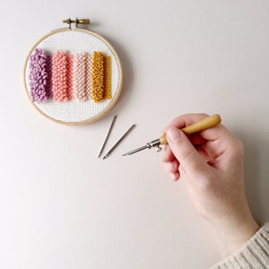May include: A close-up of a circular embroidery hoop with a white fabric background and five colourful yarn strands. The yarn strands are in shades of pink, purple, and yellow. Three embroidery needles and a wooden yarn needle are also visible.