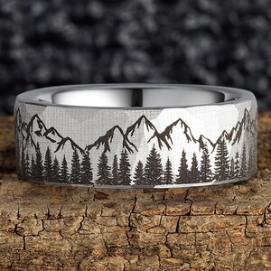May include: A silver ring with a mountain and forest design. The ring features a detailed engraving of a mountain range and evergreen trees. The ring is set against a textured wooden surface.