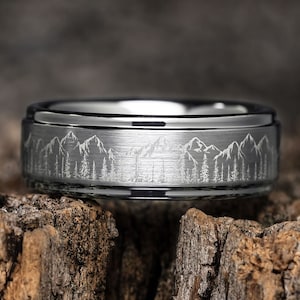 May include: A silver tungsten carbide ring with a brushed finish and an engraved mountain range design.