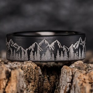 May include: A black tungsten carbide ring with a brushed finish and a mountain range and trees design engraved on the band.