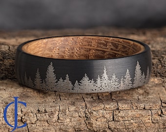 Op de afbeelding: Een zwarte ring van wolfraamcarbide met een houten inlay en een lasergegraveerd ontwerp van een bos silhouet.