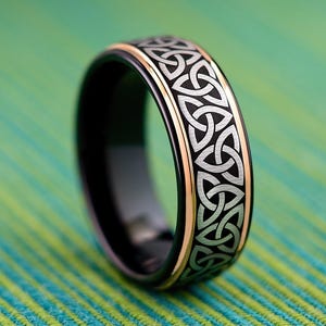 May include: A black ring with a Celtic knot design. The ring has a silver pattern on the outer band, with a gold-colored edge. The background is a green and blue striped pattern.