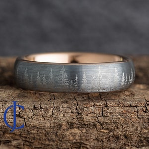 May include: A gray ring with a forest scene etched around the band. The ring has a brushed finish and a rose gold interior. The forest design features detailed tree silhouettes. The ring is displayed on a piece of wood.