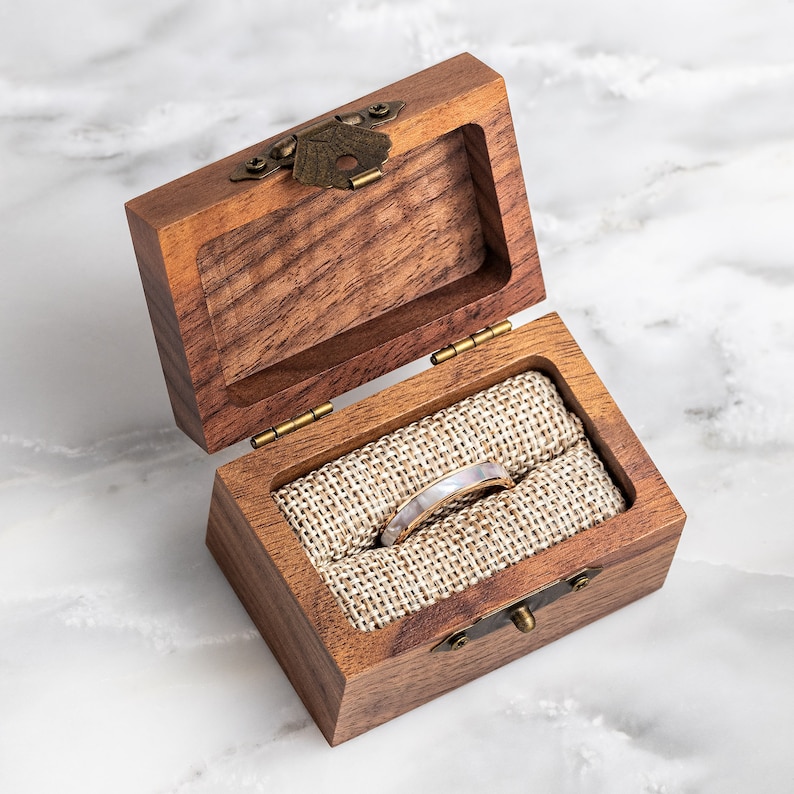 May include: A wooden ring box with a hinged lid and a brass latch. The box is lined with a beige fabric and contains a white and gold ring.