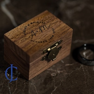 May include: A wooden ring box with a hinged lid and a brass latch. The box is engraved with the text "Ashley & Michael A+M October 17th 2021".