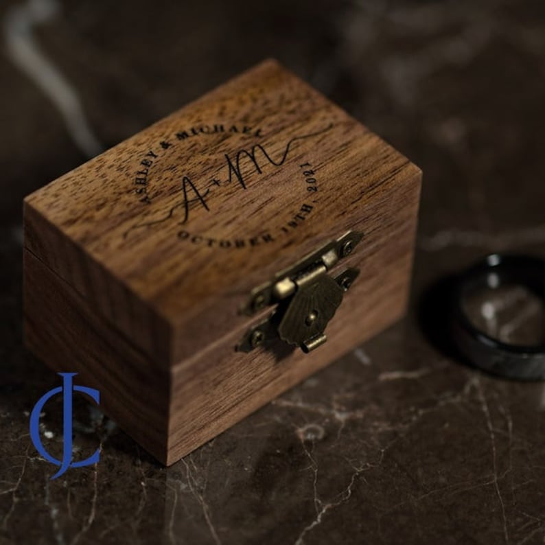 May include: A wooden ring box with a hinged lid and a brass latch. The box is engraved with the text "Ashley & Michael A+M October 17th 2021".