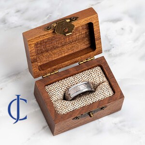Puede incluir: Un anillo de metal martillado plateado y blanco en una caja de madera con tapa abatible y cierre de lat&oacute;n.